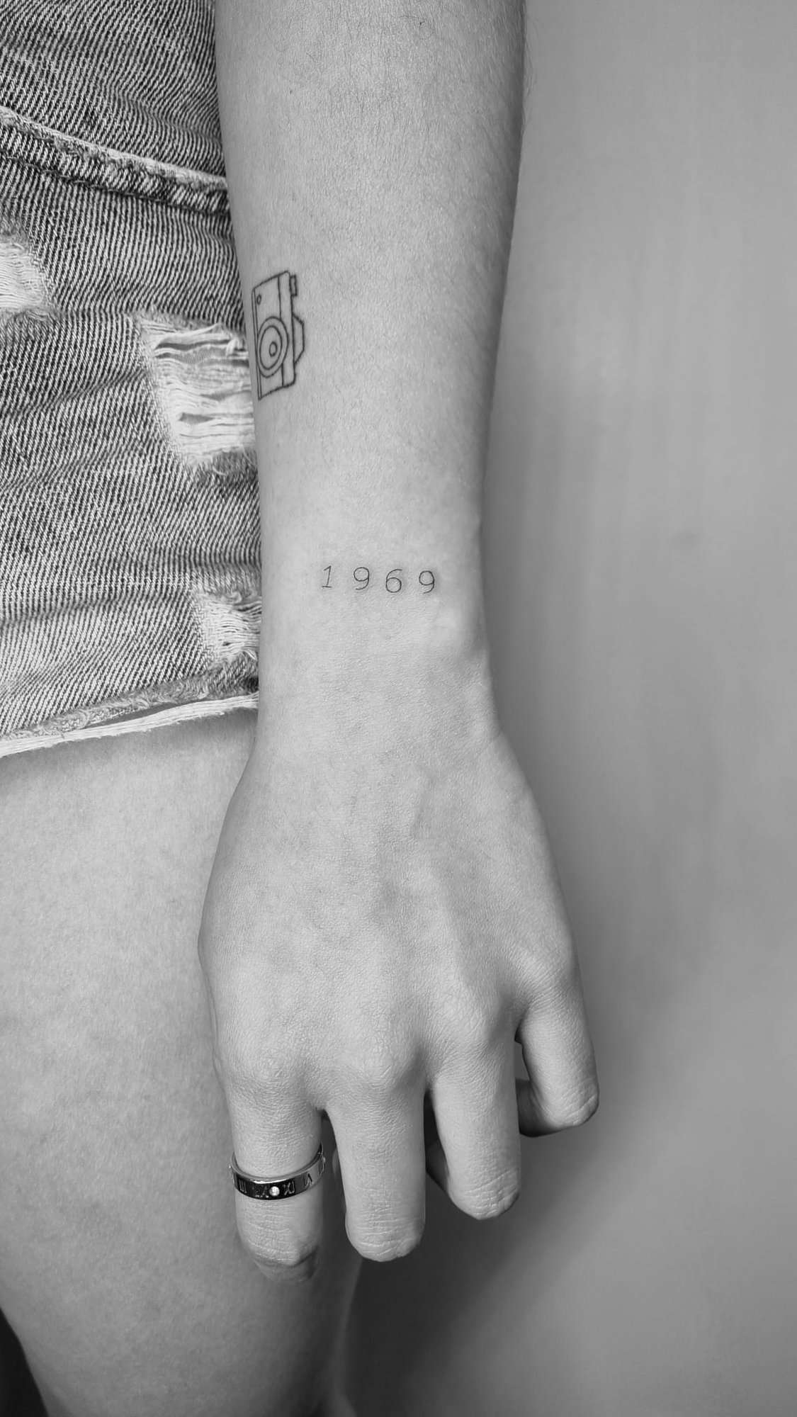 Tatuaje de camara y numero 1969 en muneca, línea fina