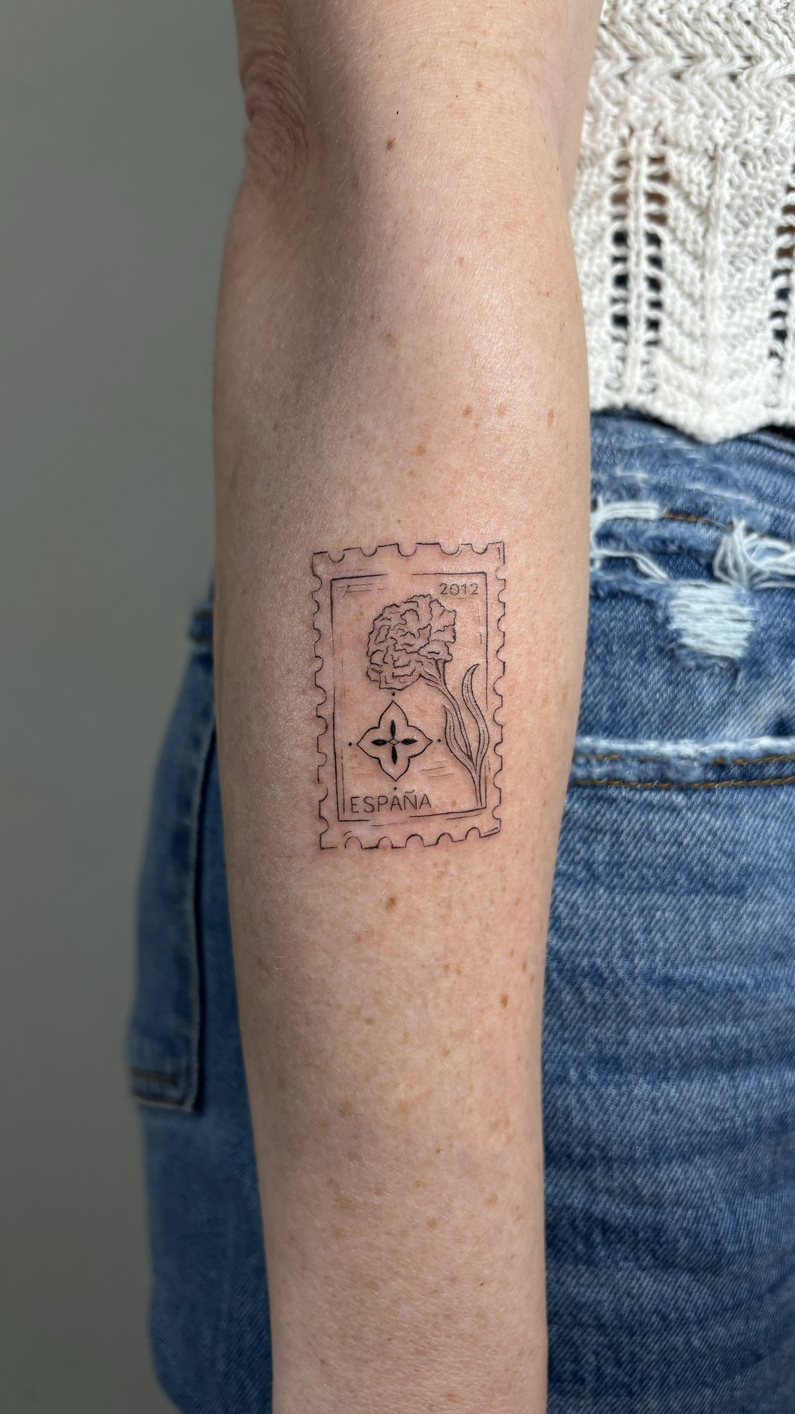 Tatuaje de sello postal espana flor en antebrazo, línea fina